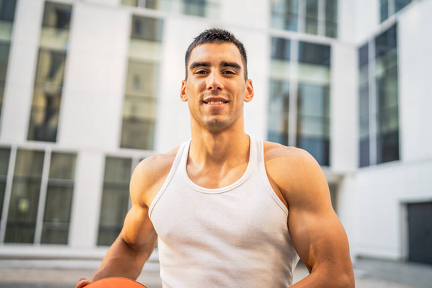 Un jeune homme caucasien athlète masculin debout extérieur tenir ballon de basket-ball porter blanc débardeur un-shirt forte musculaire personne réelle copie espace heureux sourire confiant mode de vie sain concept - Photo, image