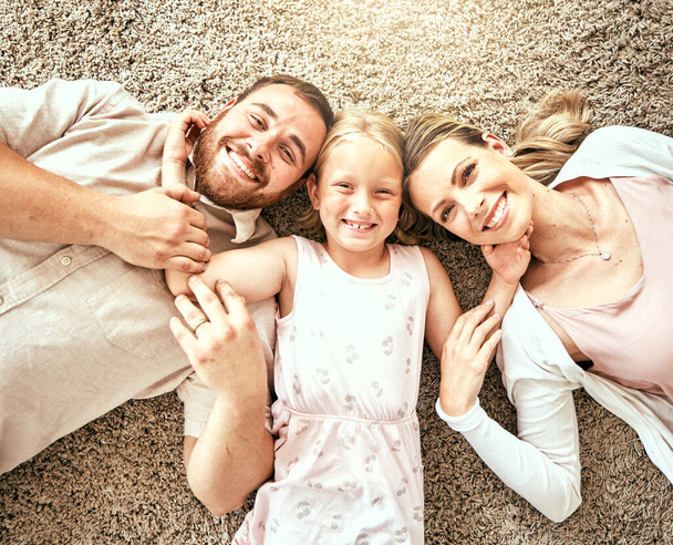 Ευτυχισμένο, πορτραίτο και παιδί με γονείς στο σαλόνι που δένουν και χαλαρώνουν μαζί στο σπίτι. Ευτυχία, αγάπη και κορίτσι ξαπλώνουν με τη μητέρα και τον πατέρα από την Αυστραλία στο πάτωμα στο σαλόνι στο σπίτι - Φωτογραφία, εικόνα