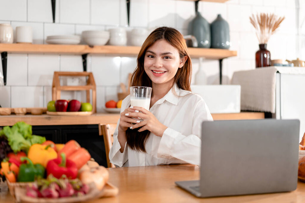 Aktivität zu Hause Freizeit Lifestylekonzept, junge Frau in Kleidung lässig in der Küche sitzen, um Film auf Laptop und Milch trinken mit genießen, während Freizeit mit Lebensstil zu Hause. - Foto, Bild