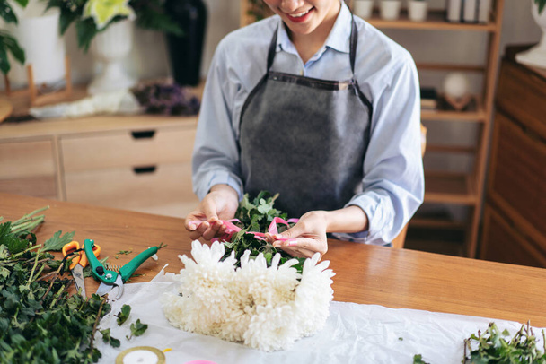 Fleuriste féminine dans le tablier en utilisant un ruban rose pour attacher pour créer et faire bouquet de fleurs de chrysanthème blanc dans sa boutique de fleurs pour la livraison au client. - Photo, image