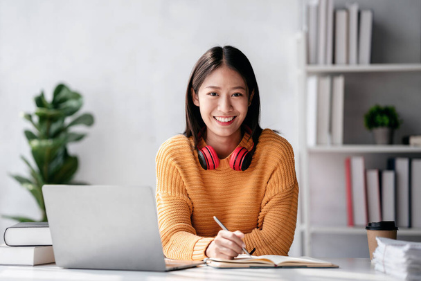 Concepto de aprendizaje a distancia, Mujer joven en suéter estudiando lección de educación en línea y escribiendo en un cuaderno mientras ve y aprende el curso a través de video en el ordenador portátil en casa. - Foto, Imagen