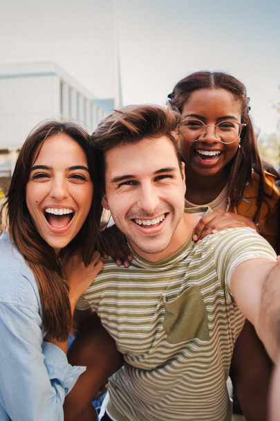 Vertical portrait of a happy multicultural friends laughing taking a selfie together. Mixed teenage cheerful students having fun and hanging out. Young multiracial joyful people looking at camera - Photo, Image