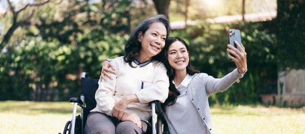 Senior en silla de ruedas y concepto de familia hija, Joven mujer asiática abrazando y selfie en el teléfono inteligente con madre discapacitada en silla de ruedas, mientras que la mujer mayor y la hija de relajación juntos en el jardín. - Foto, imagen