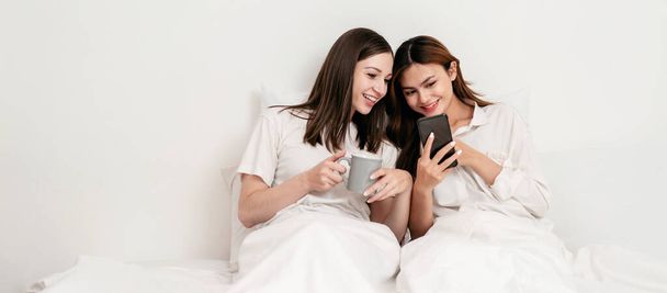 Fiatal ázsiai pár nő használ okostelefon szörfözés internet együtt és ivás forró csokoládé, miközben feküdt a takaró alatt, és támaszkodik kényelmes párnák az ágyon a hálószobában otthon. - Fotó, kép