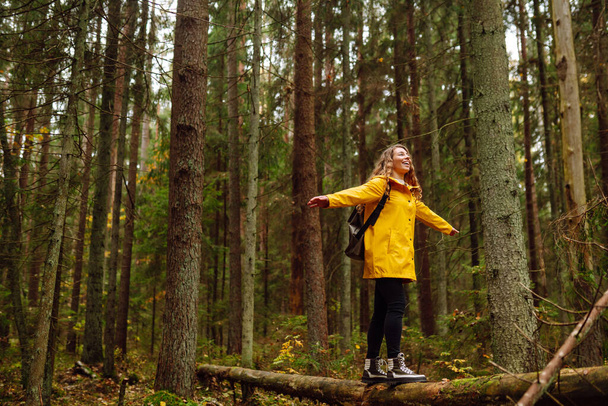 Ευτυχισμένη γυναίκα σε ένα κίτρινο παλτό απολαμβάνει το πράσινο όμορφο φυσικό δάσος γύρω της. Ένας τουρίστας με σακίδιο εξερευνά νέα μέρη, περπατώντας μέσα από το θάμνο του δάσους. - Φωτογραφία, εικόνα