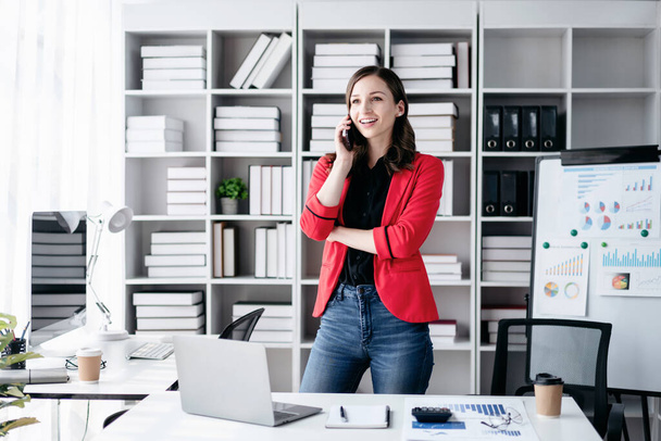 Молодая деловая женщина, используя смартфон говорить и искать бизнес-данные на ноутбуке, работая над анализом стратегии и планирования нового бизнеса в офисе. - Фото, изображение