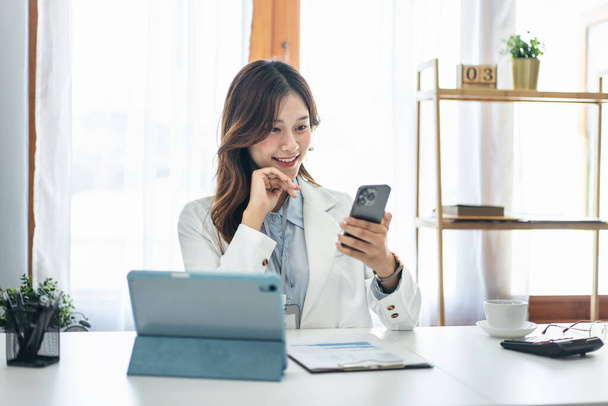 Bizneswoman w garniturze za pomocą smartfona do rozmowy z partnerami podczas czytania dokumentu biznesowego i pracy z tabletem urządzenia technologicznego w nowoczesnym biurze. - Zdjęcie, obraz