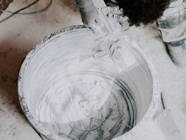 Один молодой неузнаваемый мужчина очищает лопатку от шпатлевки, держа её над пустой и грязной раковиной, вид сбоку крупным планом.. - Фото, изображение