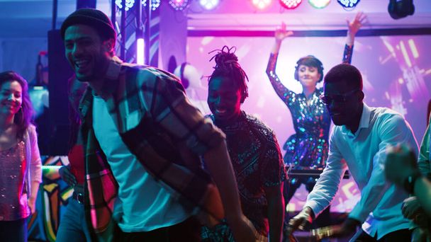 Çeşitli insanlar kulüpte parti veriyor, renkli dans pistinde canlı DJ müziğinin keyfini çıkarıyorlar. Bir grup arkadaş sahne ışıkları ve elektronik seslerle partide eğleniyor, gece kulübünde dans ediyorlar. Üçayak atışı. - Fotoğraf, Görsel