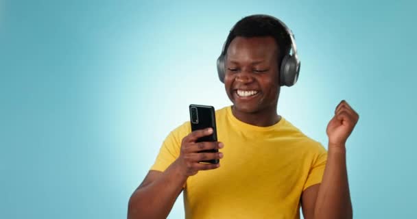 Ευτυχισμένος μαύρος άνδρας, ακουστικά και χορός στη μουσική με το τηλέφωνο σε ένα στούντιο ή μπλε φόντο. Αφρικανός άνδρας χαμογελά και απολαμβάνει ροή ήχου σε κινητό smartphone ή εφαρμογή ήχου στο mockup. - Πλάνα, βίντεο