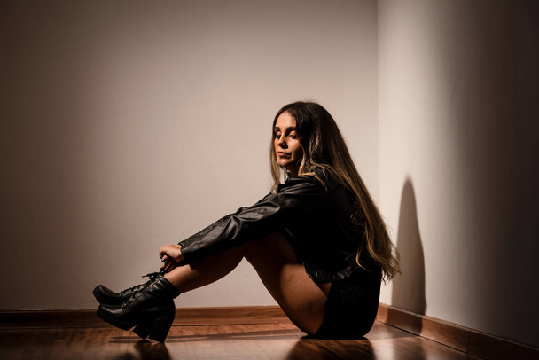 Bella giovane modella vestita di nero, abiti casual, seduta sul pavimento in legno contro la parete bianca. Ritratto studio. - Foto, immagini