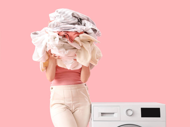 Femme avec pile de vêtements sales debout près de la machine à laver sur fond rose - Photo, image