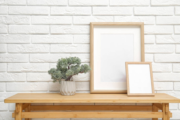 Drewniany stół z drzewem w garnku i puste ramki w pobliżu ściany z białej cegły - Zdjęcie, obraz