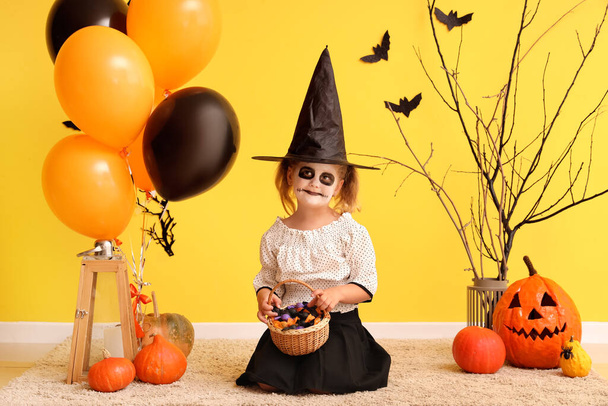 Cadılar Bayramı için sarı duvarın yanında balonlar ve şekerlerle cadı gibi giyinmiş küçük kız. - Fotoğraf, Görsel
