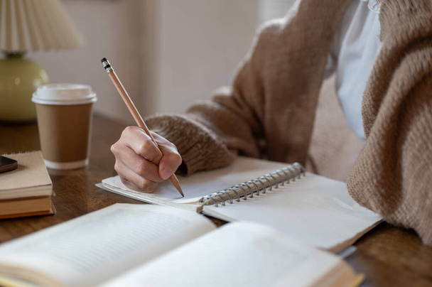 Zbliżenie kobiety w przytulnym swetrze z dzianiny prowadzącej pamiętnik, piszącej coś na książce lub odrabiającej lekcje przy stole w pomieszczeniu. - Zdjęcie, obraz