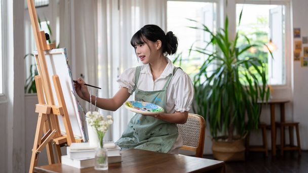 A beautiful and talented young Asian female artist painting on a canvas with water colors, working in her cosy minimalist studio alone. Creativity and inspiration concepts - Photo, Image