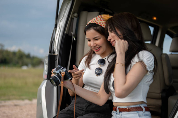 İki güzel ve mutlu Asyalı genç bayan en iyi arkadaş bir arabanın bagajında otururken konuşmaktan keyif alıyorlar, kırsala yaptıkları tatil gezisinde eğleniyorlar.. - Fotoğraf, Görsel