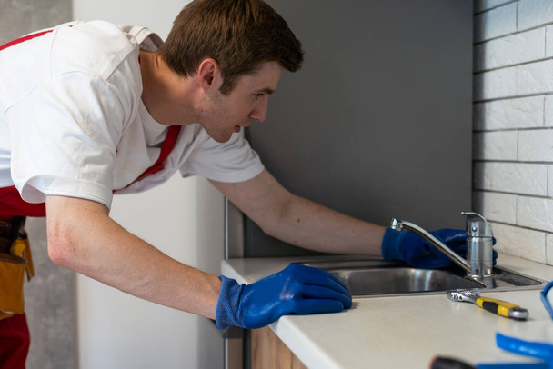 Молодой ремонтник устанавливает кран кухонной раковины в комнате кухни
. - Фото, изображение
