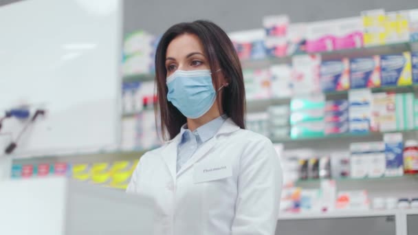 Female pharmacist looking at computer. Woman pharmacist working with cash register. Druggist wearing white robe and protective mask. Showcase with pills and syrups. Caucasian pharmacist in pharmacy. - Footage, Video