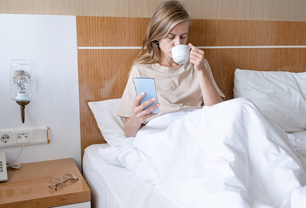 Mujer usando smartphone y tomando café tumbado en la cama en la habitación del hotel - Foto, imagen
