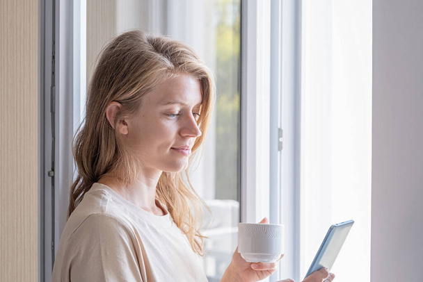 nő áll az ablak mellett, kezében mobiltelefon iszik kávét - Fotó, kép