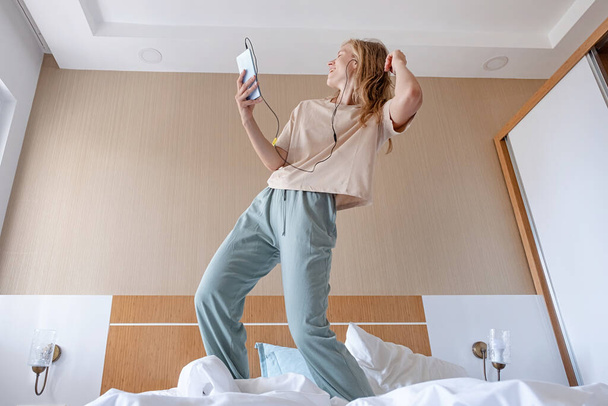 Ganzkörperporträt einer aufgeregten, glücklichen jungen Frau, die Pyjama trägt und zur Lieblingsmusik auf dem Bett im Hotelzimmer tanzt - Foto, Bild