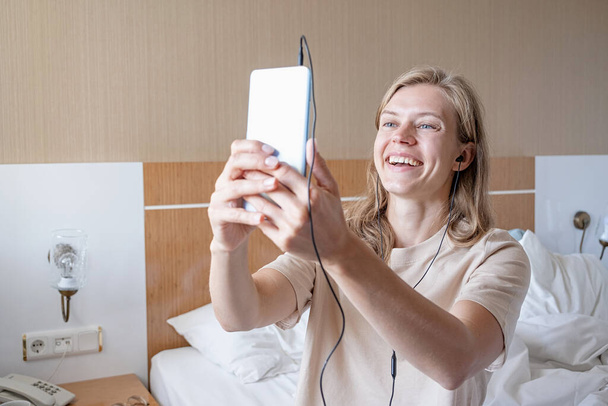 boldog nő használ fülhallgató és okostelefon ül az ágyon a szállodai szobában, bemutatja szoba barátjának videofelvételen - Fotó, kép