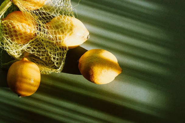 Keltaiset kypsät sitruunat putoavat muovipussista, joka jää vihreälle pöydälle pitkiksi lävistäjiksi. Trendikäs tyylikäs moderni ruoka tausta. Nettoostoskassi sitrushedelmillä aurinkoisena kesäpäivänä. Sitruunan ainesosa - Valokuva, kuva