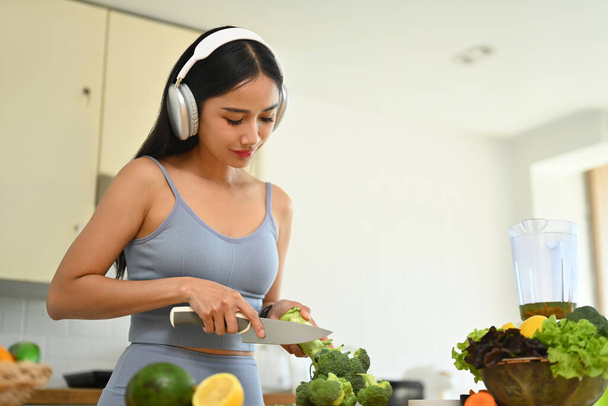 Belle femme en tenue de sport coupant le brocoli tout en préparant des aliments sains et végétariens dans la cuisine. - Photo, image