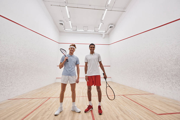 feliz interracial macho amigos en sportswear mirando cámara y sosteniendo raquetas para squash - Foto, Imagen