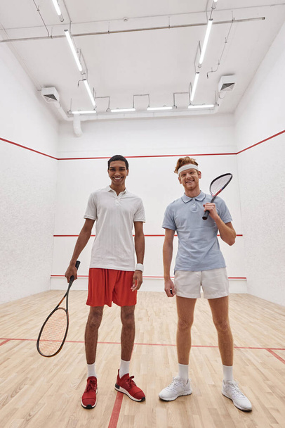 vertical shot, happy interracial male friends looking at camera and holding racquets for squash - Photo, Image
