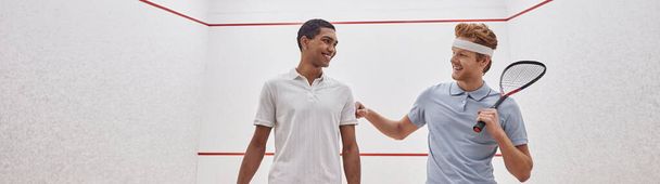 feliz interracial los hombres en sportswear sonriendo y sosteniendo raquetas para squash dentro de la corte, bandera - Foto, imagen