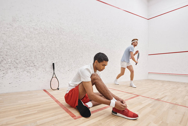 Afrikaans amerikaanse speler strikken schoenveters terwijl zitten in de buurt roodharige vriend in van squash court - Foto, afbeelding