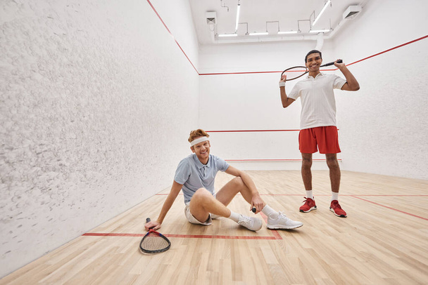happy man holding racquet while sitting near african american friend inside of squash court - Photo, Image