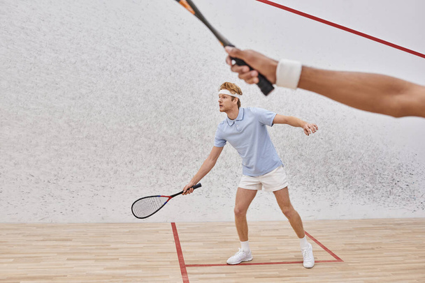 redhead man in sportswear and headband playing with african american friend inside of squash court - Foto, Imagen