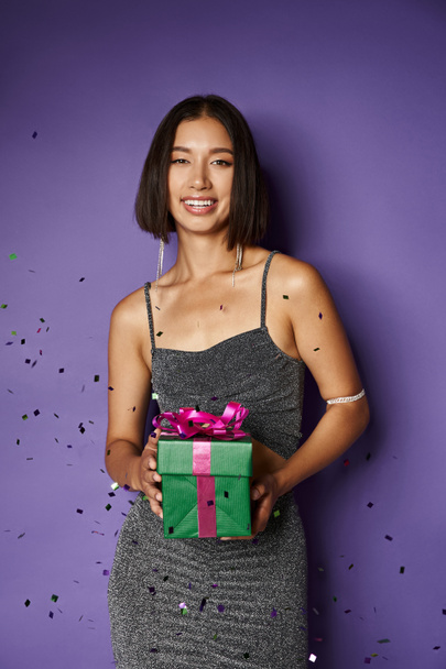 rayonnante asiatique femme en partie robe tenant enveloppé cadeau de Noël près de tomber confettis sur violet - Photo, image