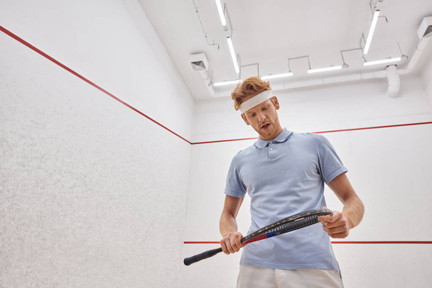 tired man in headband and sportswear breathing heavily and looking at squash racquet inside of court - Zdjęcie, obraz