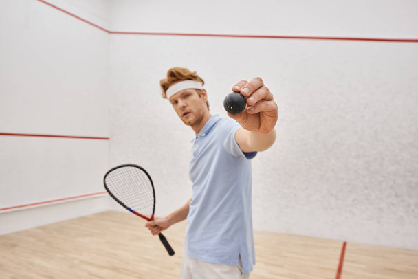 redhead man in sportswear showing squash ball and holding racquet while standing inside of court - Foto, Imagen