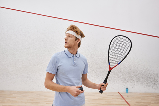 rossa uomo in abbigliamento sportivo in possesso di palla da squash e racchetta mentre in piedi all'interno della corte coperta - Foto, immagini