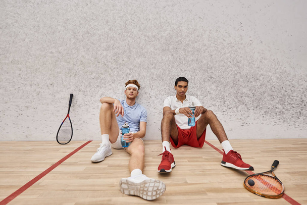 due amici interrazziali stanchi con racchette da squash e bottiglia con acqua seduta sul pavimento in tribunale - Foto, immagini