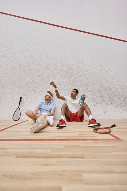 два межрасовых спортсмена с ракетками для сквоша и бутылкой с водой, сидящей на полу в суде - Фото, изображение