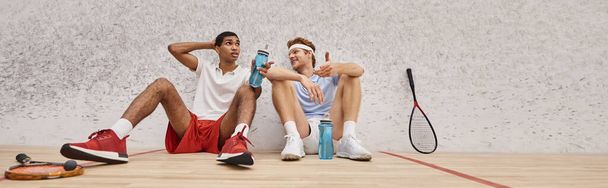 striscione, sportivi interrazziali con racchette da squash e bottiglie con acqua seduti sul pavimento in tribunale - Foto, immagini