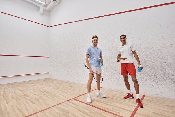 cheerful interracial sportsmen with squash racquets and bottles with water standing inside of court - Photo, image