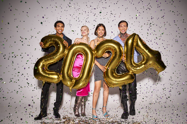 joyful multiethnic friends holding balloons with 2024 numbers near falling confetti, New Year party - Fotografie, Obrázek