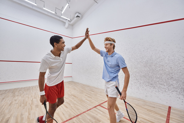 Spor giyim sektöründe gülümseyen çok kültürlü arkadaşlar mahkemede squash oynadıktan sonra beşlik çakıyor. - Fotoğraf, Görsel