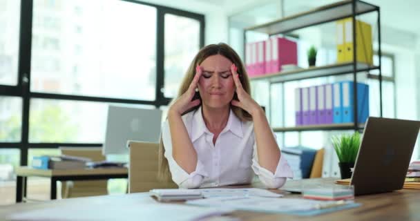 Joven mujer frustrada que trabaja en el escritorio de la oficina delante de la computadora portátil y que sufre de dolores de cabeza diarios crónicos. Estrés por exceso de trabajo y migraña en el trabajo - Metraje, vídeo