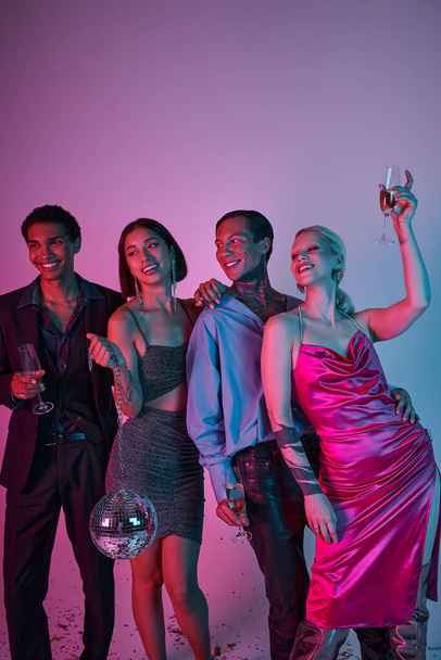 joyful interracial friends holding champagne glasses and disco ball on purple pink, Christmas party - Photo, Image