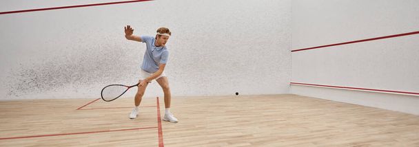 joven y activo pelirrojo en ropa deportiva jugando squash dentro de la corte, desafío bandera - Foto, Imagen