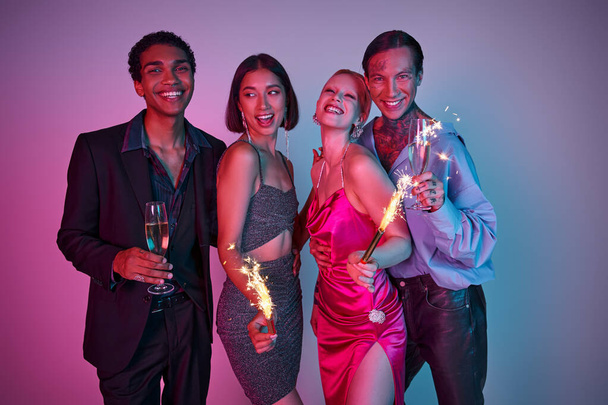 happy multiethnic friends holding sparklers and champagne, celebrating Christmas on purple pink - Photo, Image