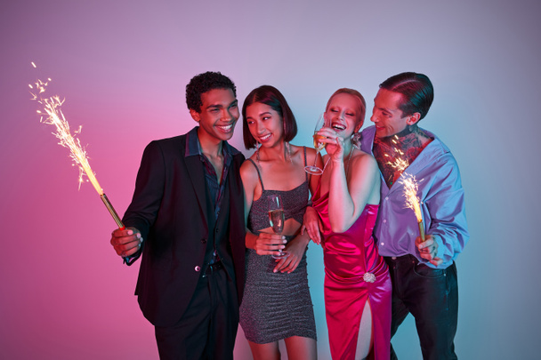 Christmas celebration, radiant multiethnic friends holding sparklers and champagne on purple pink - Foto, imagen